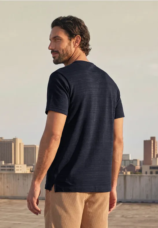 Ein Mann mit dunkelblauer T-Shirt und beige Shorts steht auf einem Dach und blickt über die Stadt.