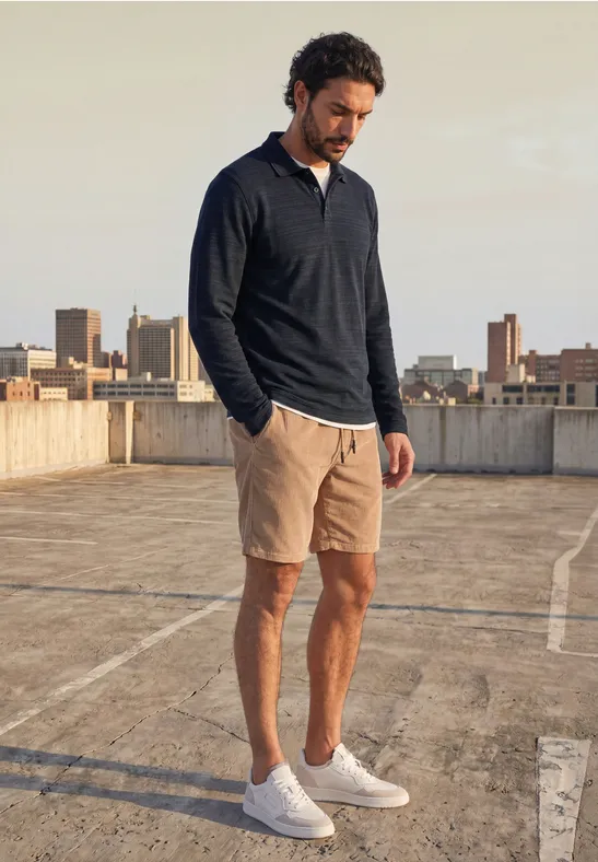 Ein Mann steht auf einem Parkdeck mit Stadtkulisse, trägt dunkelblauen Pullover, beige Shorts und weiße Sneaker.