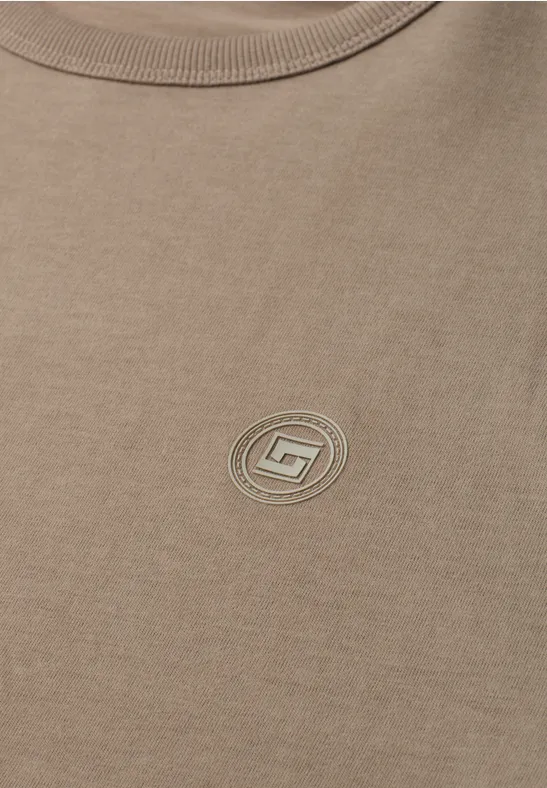 Beiges T-Shirt mit rundem, dezentem Logo mittig auf der Brust und Rundhalsausschnitt.