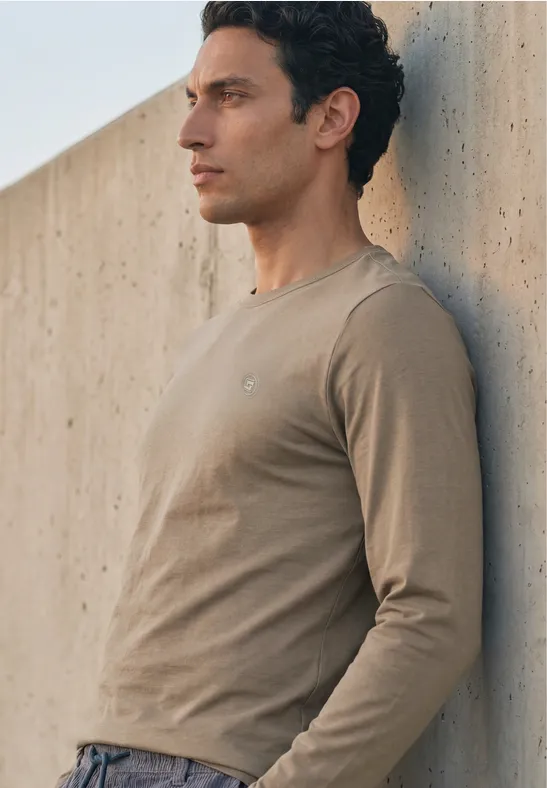 Ein junger Mann lehnt im Profil an einer Betonwand und trägt ein beiges Langarmshirt.