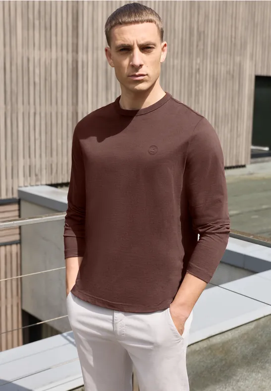 Ein Mann in braunem Langarmshirt und weißen Hosen steht entspannt auf einer Dachterrasse.