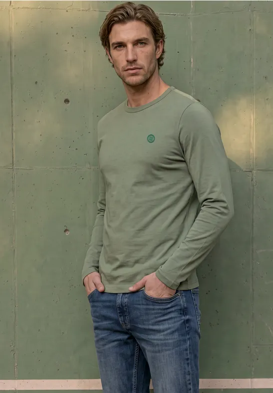 Ein junger Mann mit hellbraunen Haaren trägt ein grünes Langarmshirt und Jeans vor einer grünen Wand.