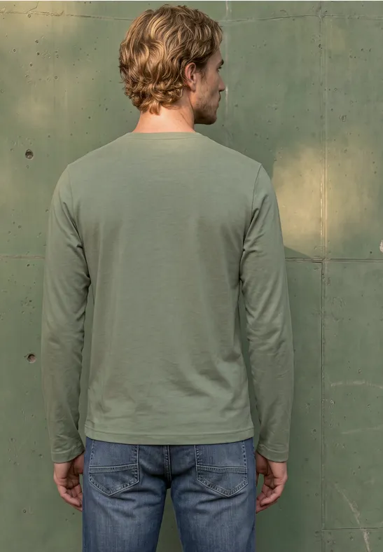 Ein Mann von hinten in legerem, grünen Langarmshirt und Jeans steht vor einer grünen Wand.