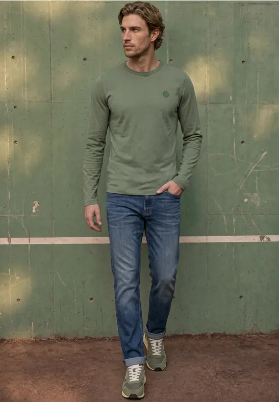 Ein Mann in grünem Langarmshirt, Jeans und Sneakers posiert vor einer grünen Wand auf einem Tennisplatz.