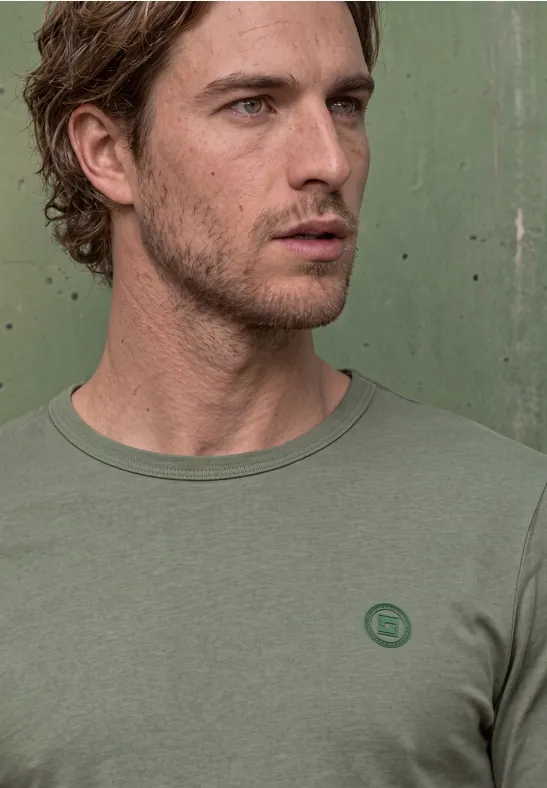 Ein junger Mann mit leicht gewelltem braunem Haar trägt ein schlichtes grünes T-Shirt vor einer grünen Wand.