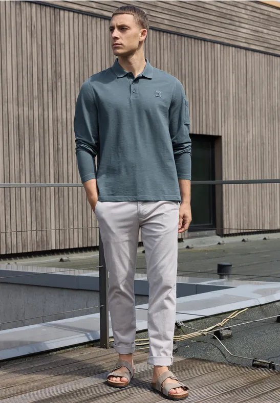 Mann in petrolfarbenem Langarm-Poloshirt, hellen Chinos und Sandalen steht auf einem Dach vor Holzfassade.