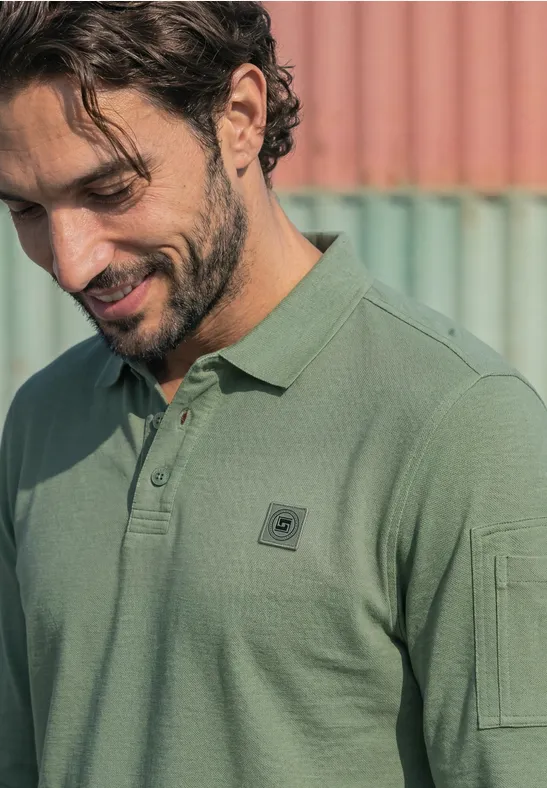 Ein junger Mann in grünem Poloshirt lächelt, den Blick nach unten gerichtet vor pastellfarbenem Hintergrund.