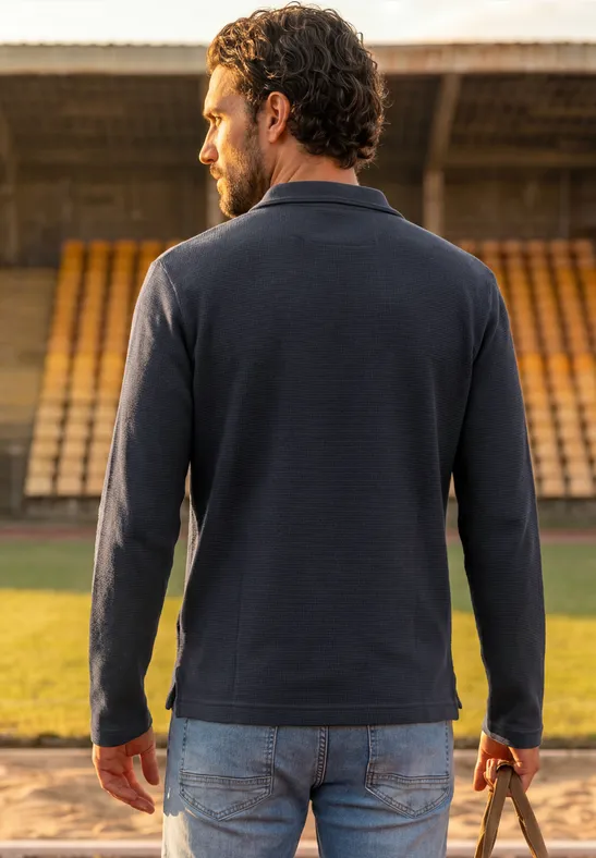 Ein Mann trägt einen dunklen Pullover und hält einen Ball auf einer Stadiontribüne.