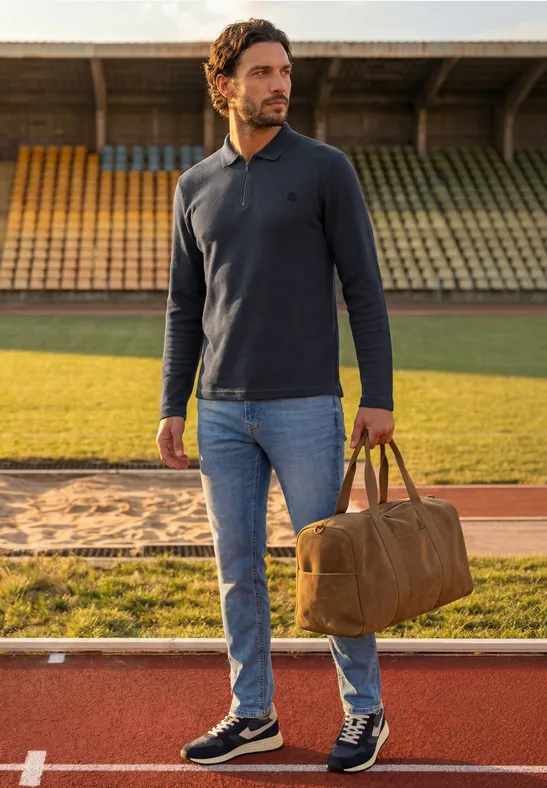 Mann mit Cap, blauem Langarm-Polo, Jeans und Sneakers trägt Tasche auf Bahn im Stadion