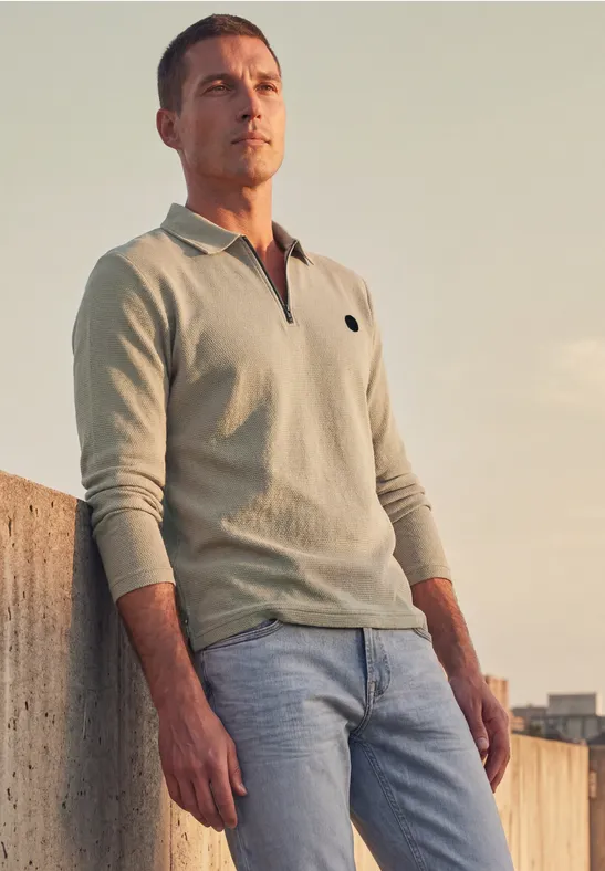 Ein Mann lehnt an einer Mauer, trägt helles Poloshirt und Jeans und blickt im Abendlicht in die Ferne.