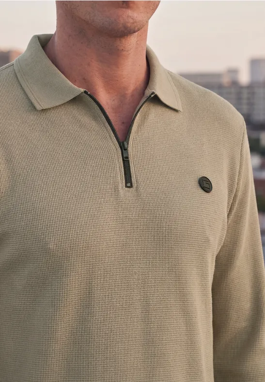Ein Mann trägt ein khakifarbenes Strick-Poloshirt mit halbem Reißverschluss und kleinem Logo auf der Brust.