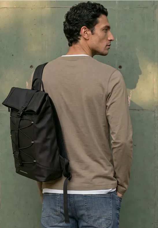 Ein Mann mit schwarzem Rucksack, trägt ein langärmeliges Shirt und Jeans, steht seitlich vor einer grünen Wand