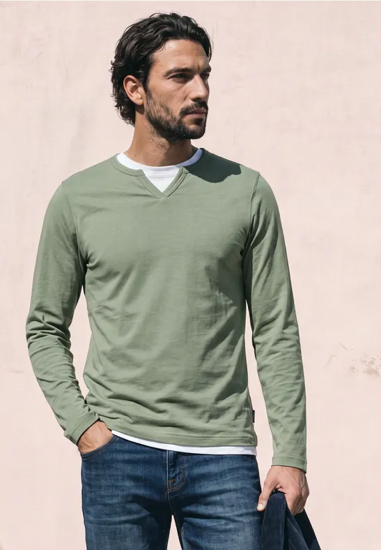 Een man met groen shirt met lange mouwen en spijkerbroek staat voor een lichte muur en kijkt opzij.