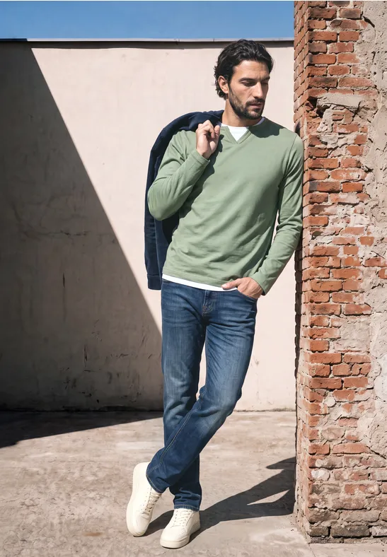 Man leunt tegen een bakstenen muur in een zonnige binnenplaats, draagt jeans, groen trui, jas over schouder en kijkt weg.