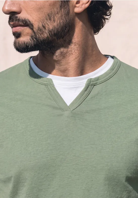 Een man met een korte zwarte baard draagt een lichtgroene trui over een wit T-shirt en kijkt opzij.