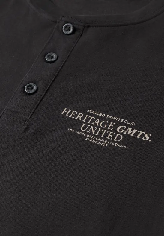 T-shirt noir avec patte de boutonnage et inscription dorée 'Heritage GMTS UNITED'.