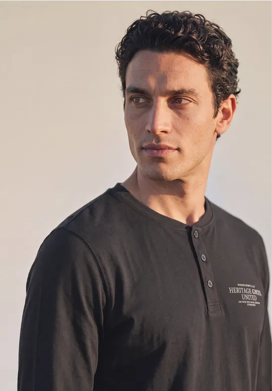 Un homme aux cheveux foncés en T-shirt noir type Henley regarde vers la gauche sur fond clair.
