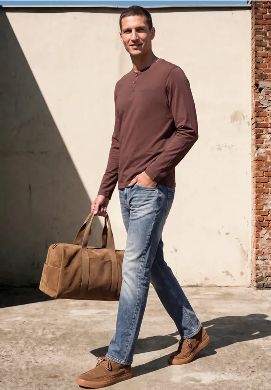 Un homme en t-shirt marron, jean et chaussures brunes porte un sac en toile et sourit en marchant.