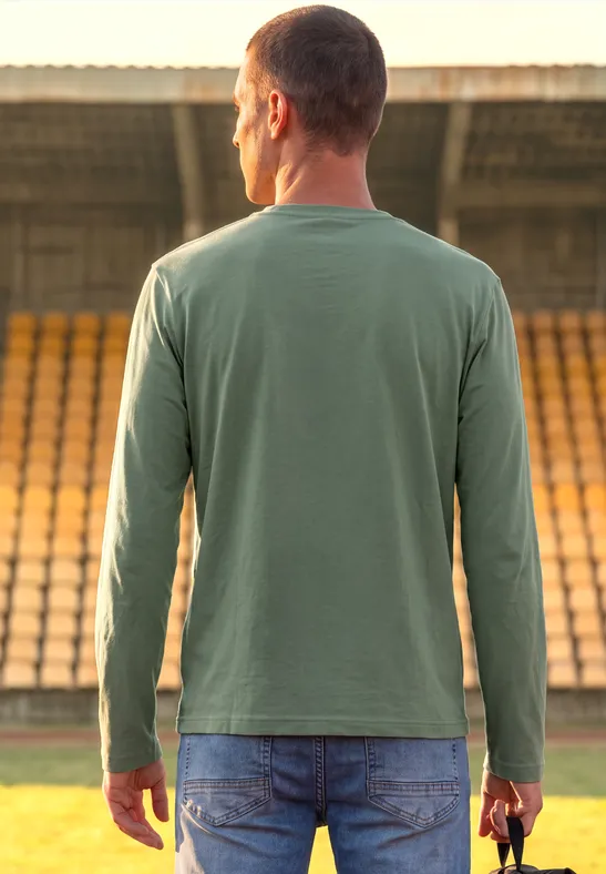 Un homme en jean et t-shirt vert se tient dans un stade en tenant un casque de football américain rouge.