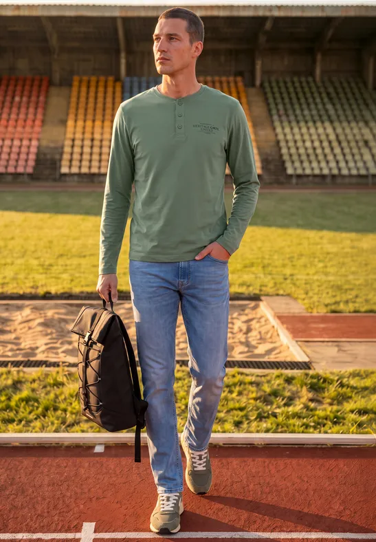 Un homme en t-shirt à manches longues vert et jean porte un sac à dos noir sur une piste.