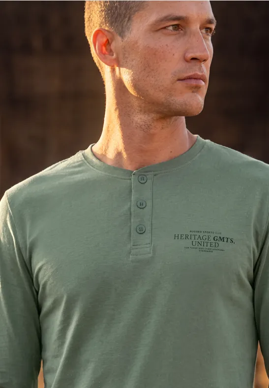 Un jeune homme aux cheveux courts porte un t-shirt vert olive à manches longues avec patte de boutonnage et inscription.