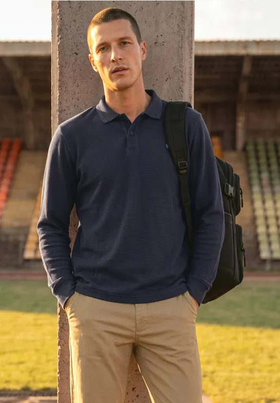Ein Mann in blauem Poloshirt und beigen Hosen steht mit Rucksack vor leeren Tribünen.