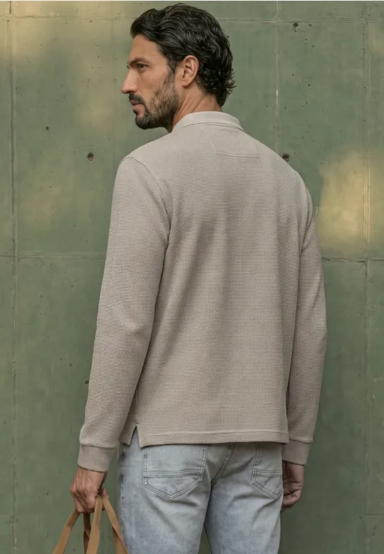 Ein Mann in beigem Langarm-Poloshirt und Jeans steht seitlich vor grünem Hintergrund und hält eine Tasche.
