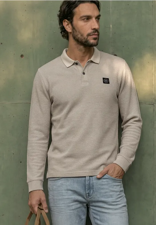 Junger Mann mit dunklem Haar und Bart trägt hellbeiges Poloshirt und Jeans, lehnt an einer grünen Wand.