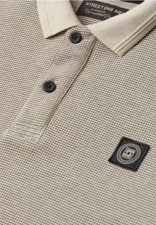 Beige Waffel-Polo mit khakifarbenem Kragen, schwarzen Knöpfen und quadratischem Logo-Patch auf der Brust.