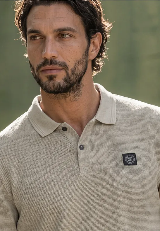 Ein dunkelhaariger, bärtiger Mann in einem beigen Poloshirt mit kleinem Logo auf der Brust.