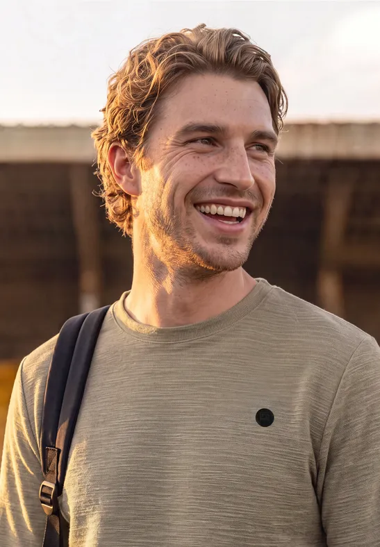 Ein junger Mann in einem beigen T-Shirt mit Rucksackschultergurt lächelt im Freien.