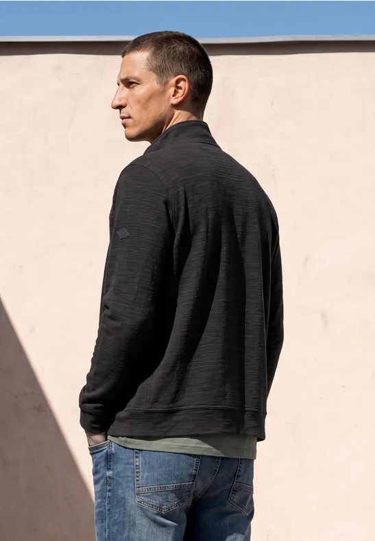 Ein Mann in Jeans und dunklem Pullover steht vor einer hellen Wand, sein Rücken dem Betrachter zugewandt.