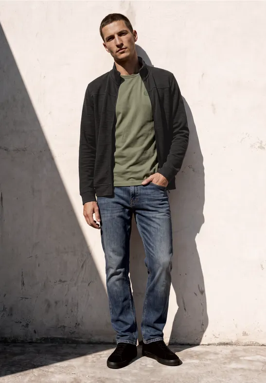 Ein Mann steht vor einer hellen Wand, trägt Jeans, grünen Pullover, schwarze Jacke und blickt entspannt geradeaus.