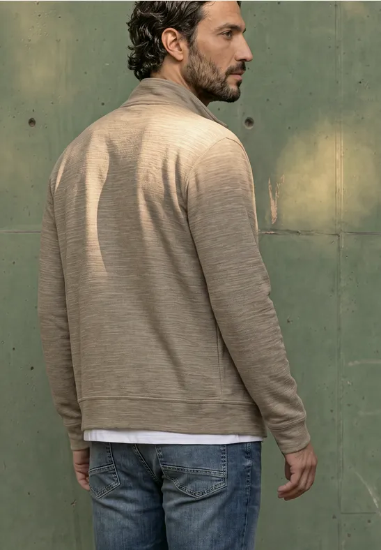 Ein Mann steht mit dem Rücken zur Kamera, trägt beiges Cardigan, weißes T-Shirt und Jeans vor grüner Wand.