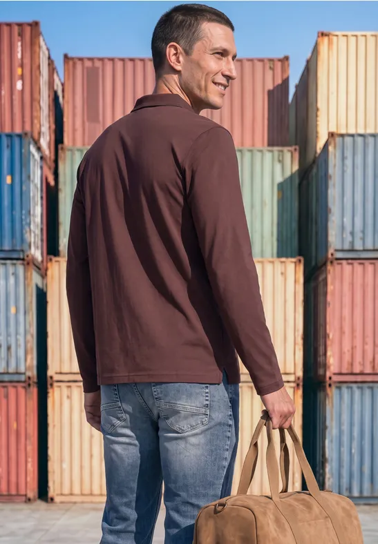 Ein Mann in braunem Hemd und Jeans trägt eine Tasche vor bunten Transportcontainern.