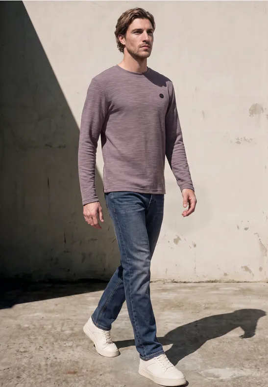 Ein Mann in Jeans, grauem Pullover und weißen Schuhen geht bei Sonne an einer Wand vorbei.