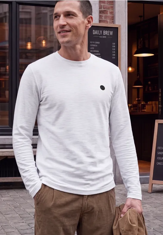 Ein Mann in weißem Langarmshirt und brauner Hose steht vor einem Café und lächelt leicht.
