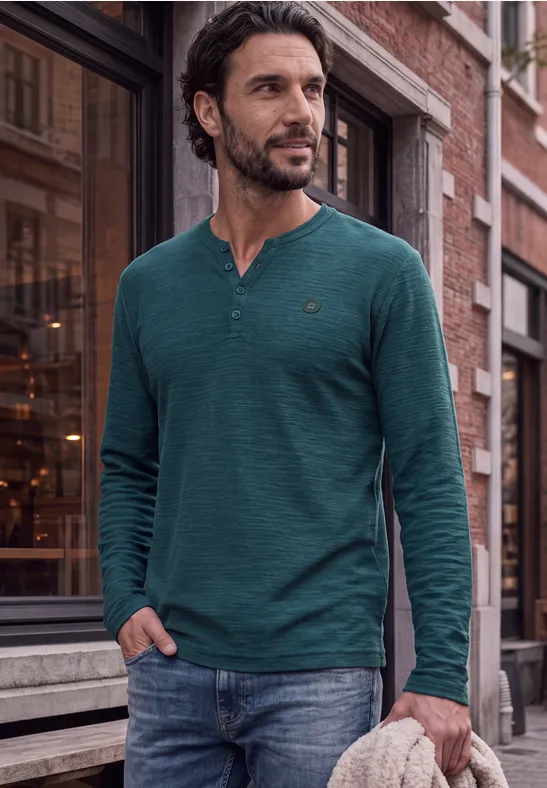 Een man draagt een groen shirt met lange mouwen en jeans en staat op een stadsstraat.