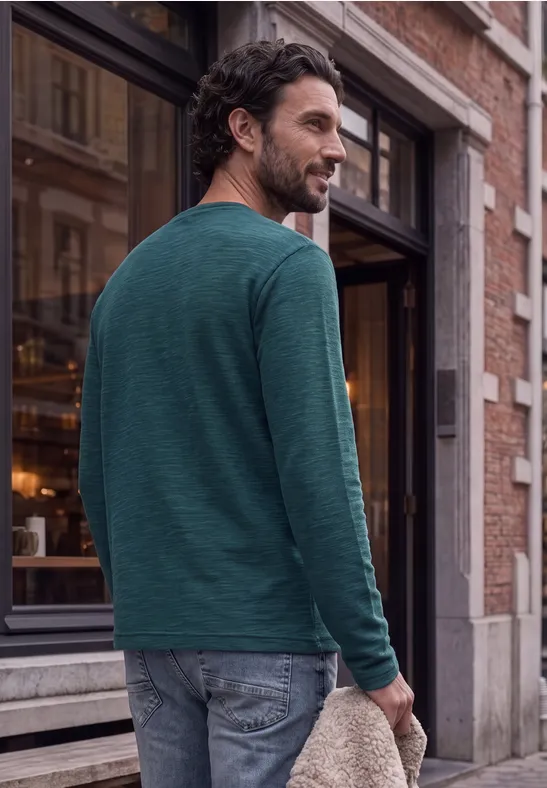 Een man in een groen shirt met lange mouwen en spijkerbroek loopt langs een gebouw en kijkt lachend over zijn schouder.