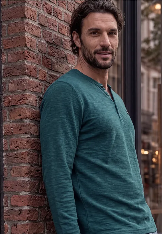 Een bebaarde man in een groen shirt met lange mouwen leunt ontspannen tegen een bakstenen muur in de stad.
