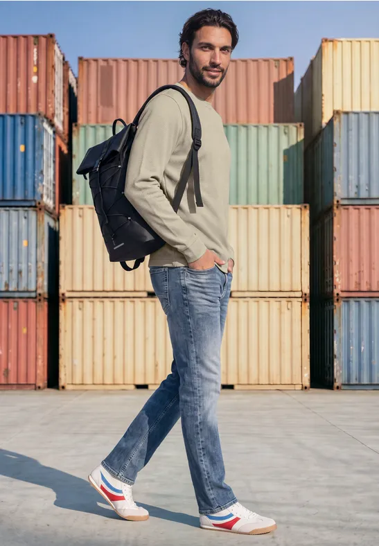 Een man in jeans, witte sneakers en rugzak staat zijwaarts voor kleurrijke containers.