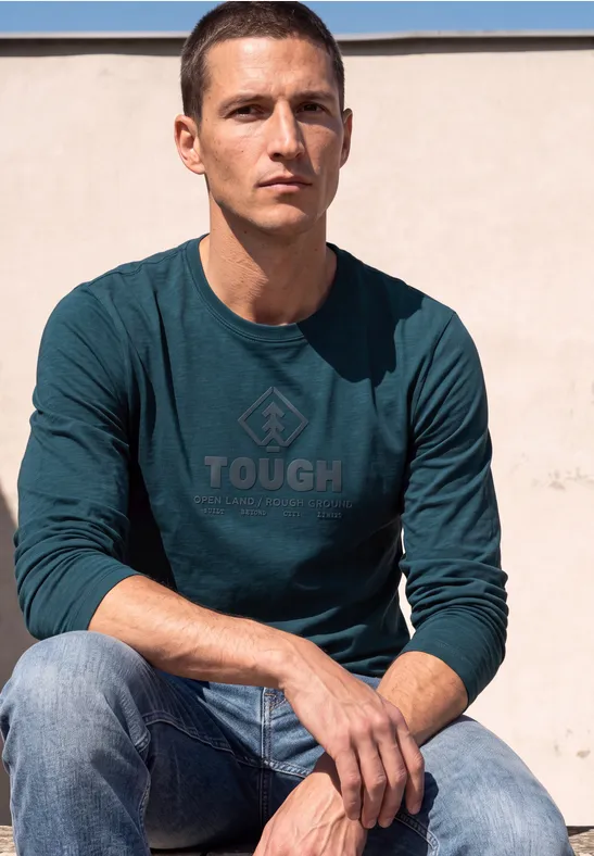 Ein Mann sitzt vor einer Wand, trägt Jeans und ein langärmeliges T-Shirt mit 'TOUGH'-Aufdruck.