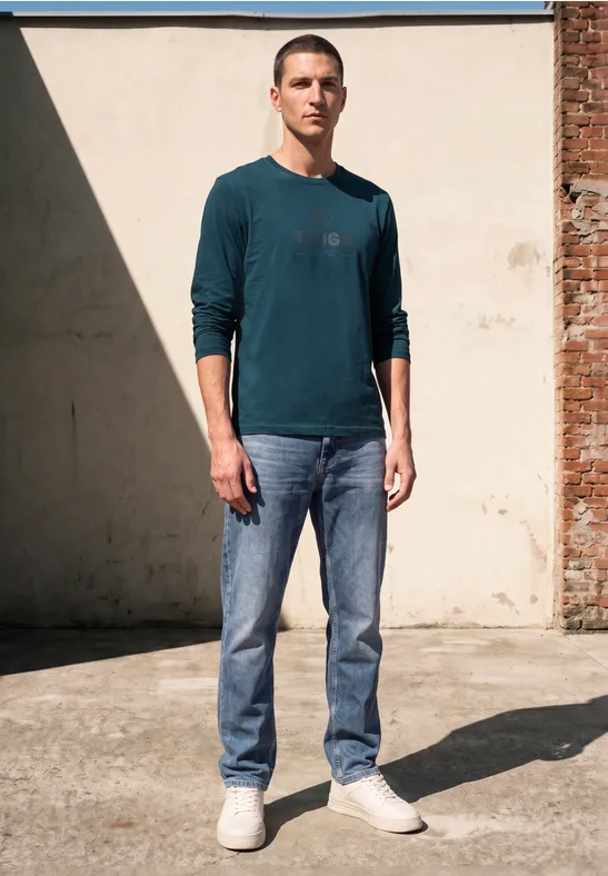 Ein Mann in einem grünen Pullover und Jeans steht selbstbewusst vor einer Wand und schaut in die Kamera.