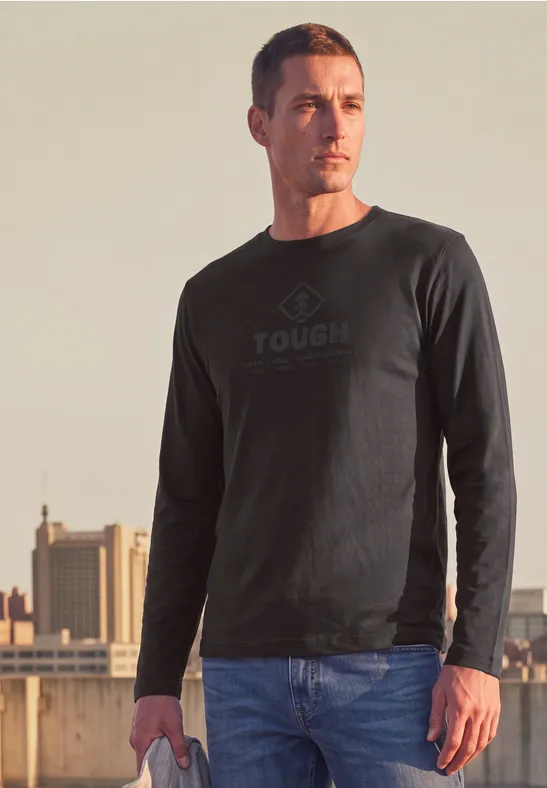 Ein Mann trägt ein schwarzes Sweatshirt mit dem Aufdruck 'TOUGH' und steht auf einem Dach vor städtischer Kulisse.
