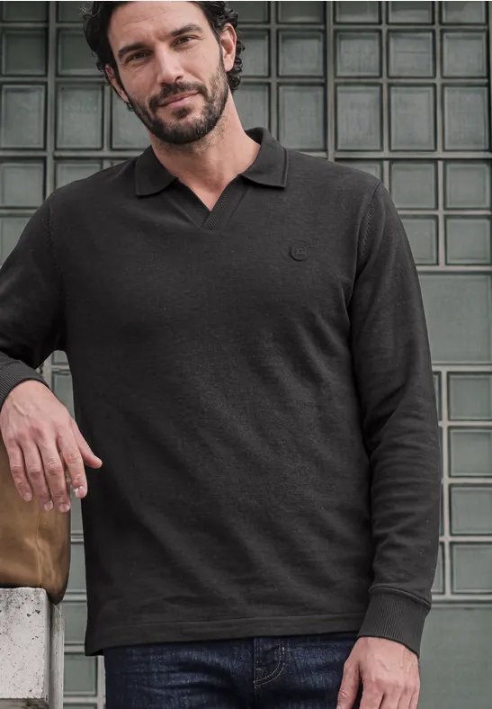 Een man leunt tegen een glasstenen muur en draagt een zwarte poloshirt met lange mouwen en jeans.