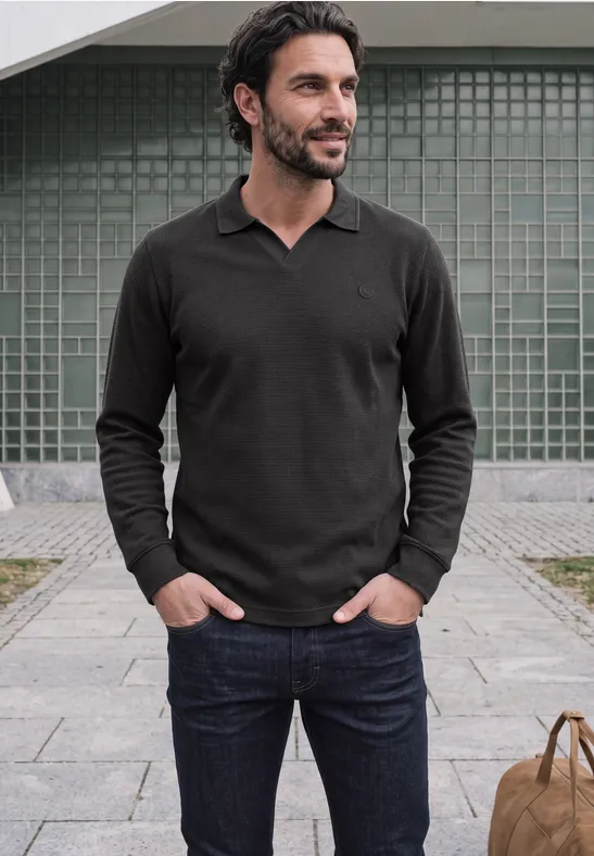 Een man in een donker poloshirt met lange mouwen staat ontspannen met handen in zakken, tas ernaast.