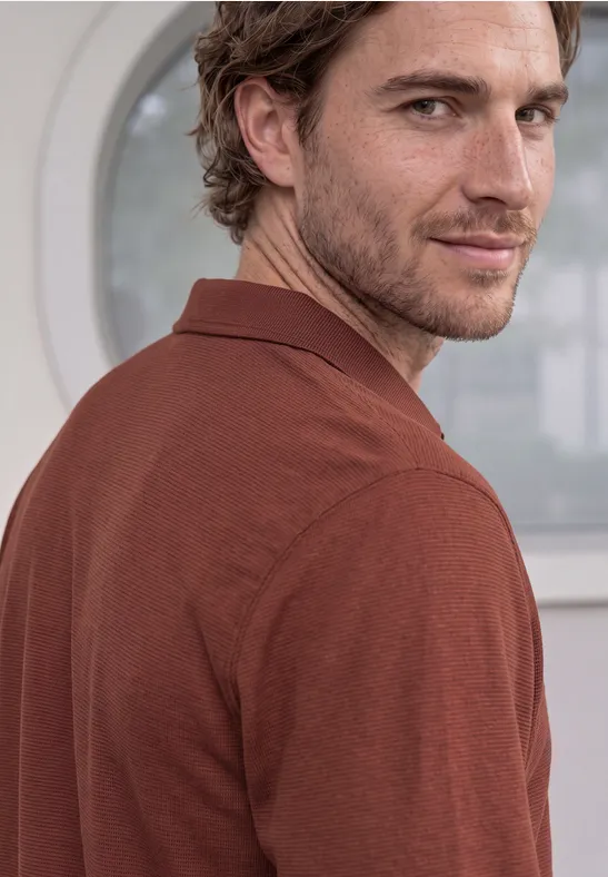 Een man in een bruin poloshirt kijkt glimlachend over zijn schouder voor een licht raam.