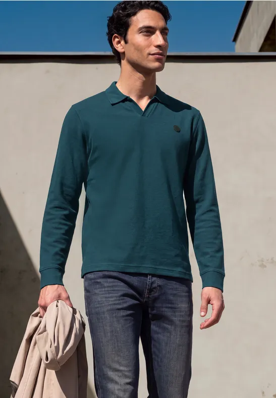 Een man draagt een donkergroen poloshirt met lange mouwen, een donkere spijkerbroek en houdt een jas vast.