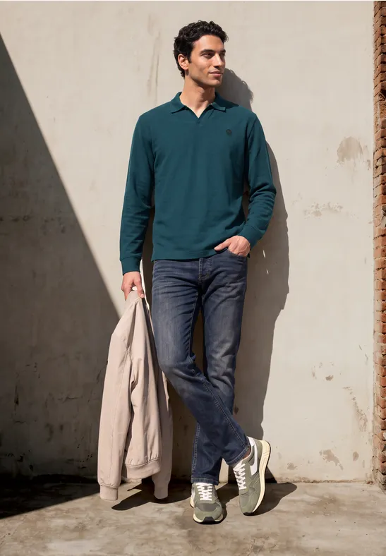 Een man leunt tegen een muur, draagt een groen shirt, jeans, sneakers en houdt een beige jas vast.