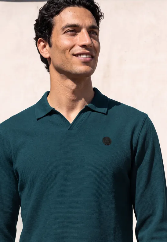 Een lachende man in een donkergroen poloshirt met lange mouwen, tegen een lichte achtergrond.
